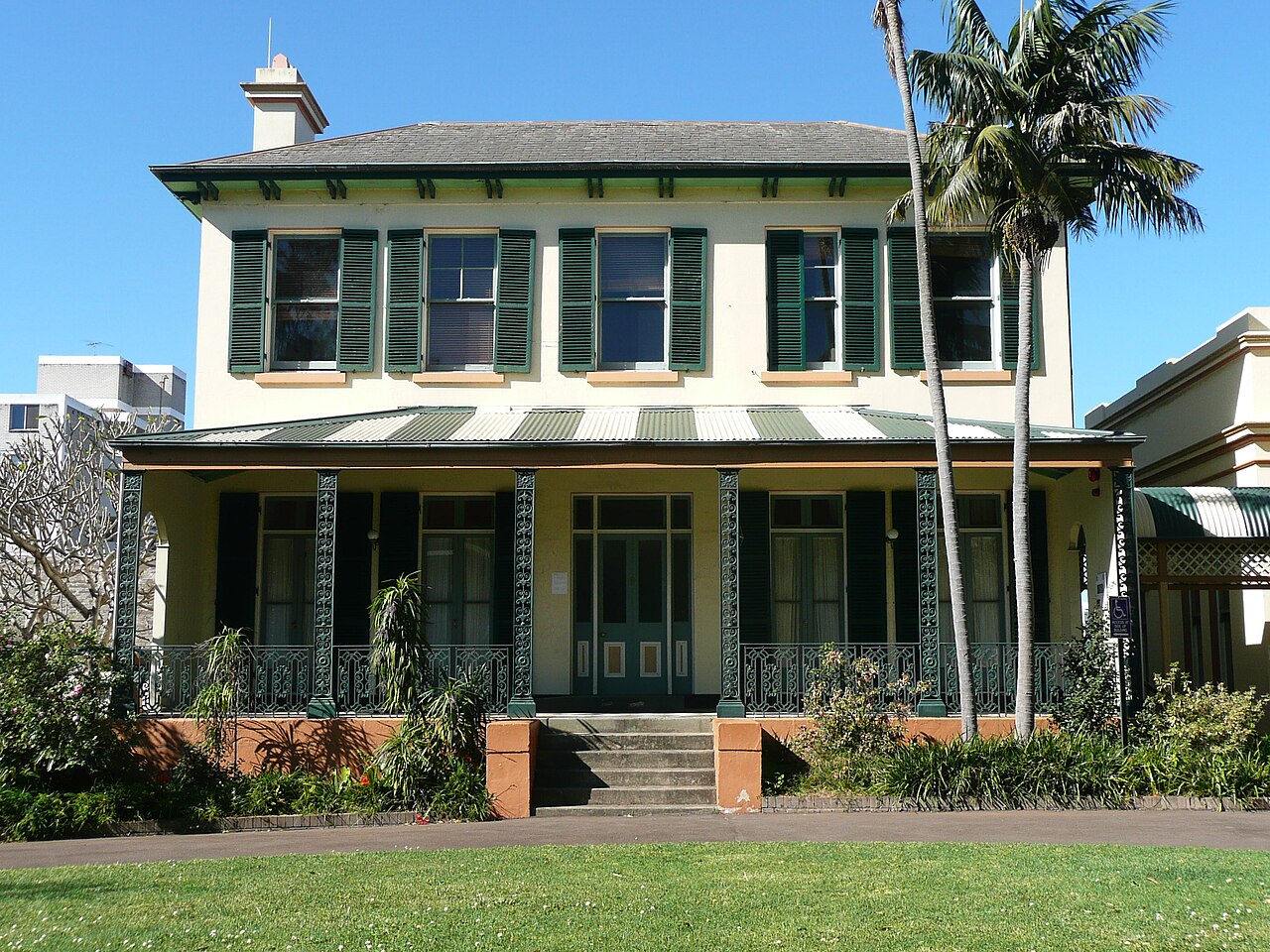 Bidura’s grand cream façade, green shutters, hipped slate roof and ironwork verandah, framed by palms and a front garden, photographed in 2009.
