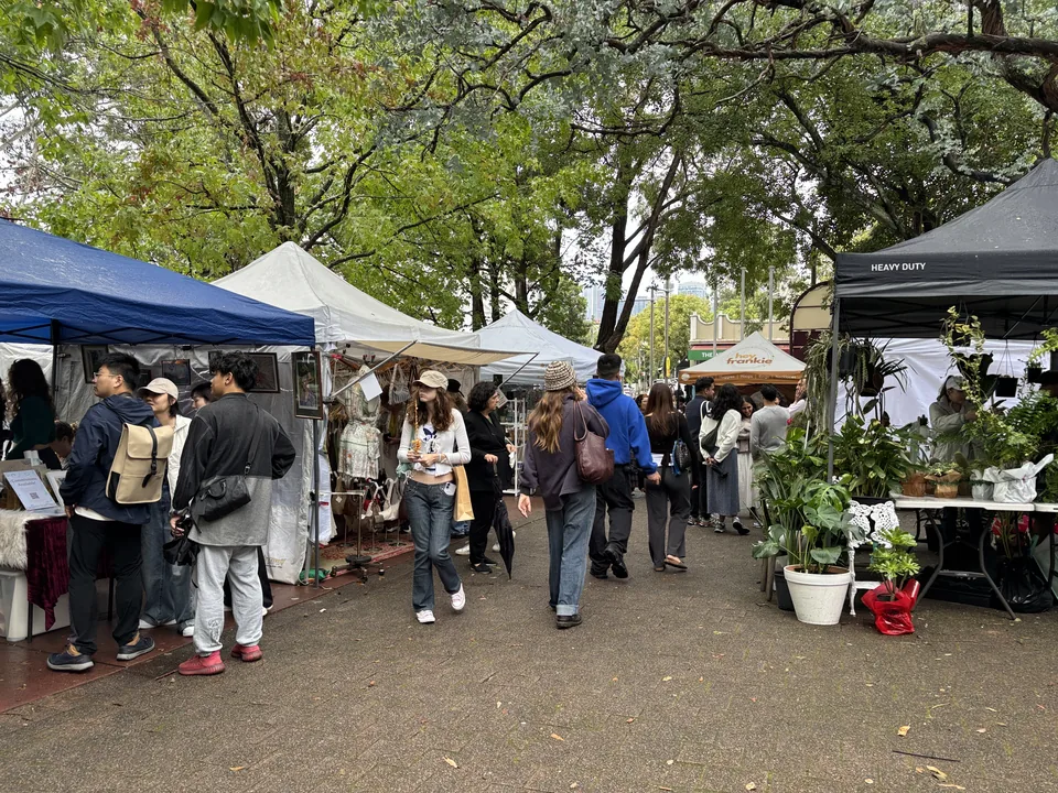 Shoppers browsing Glebe Markets stalls beneath leafy trees.